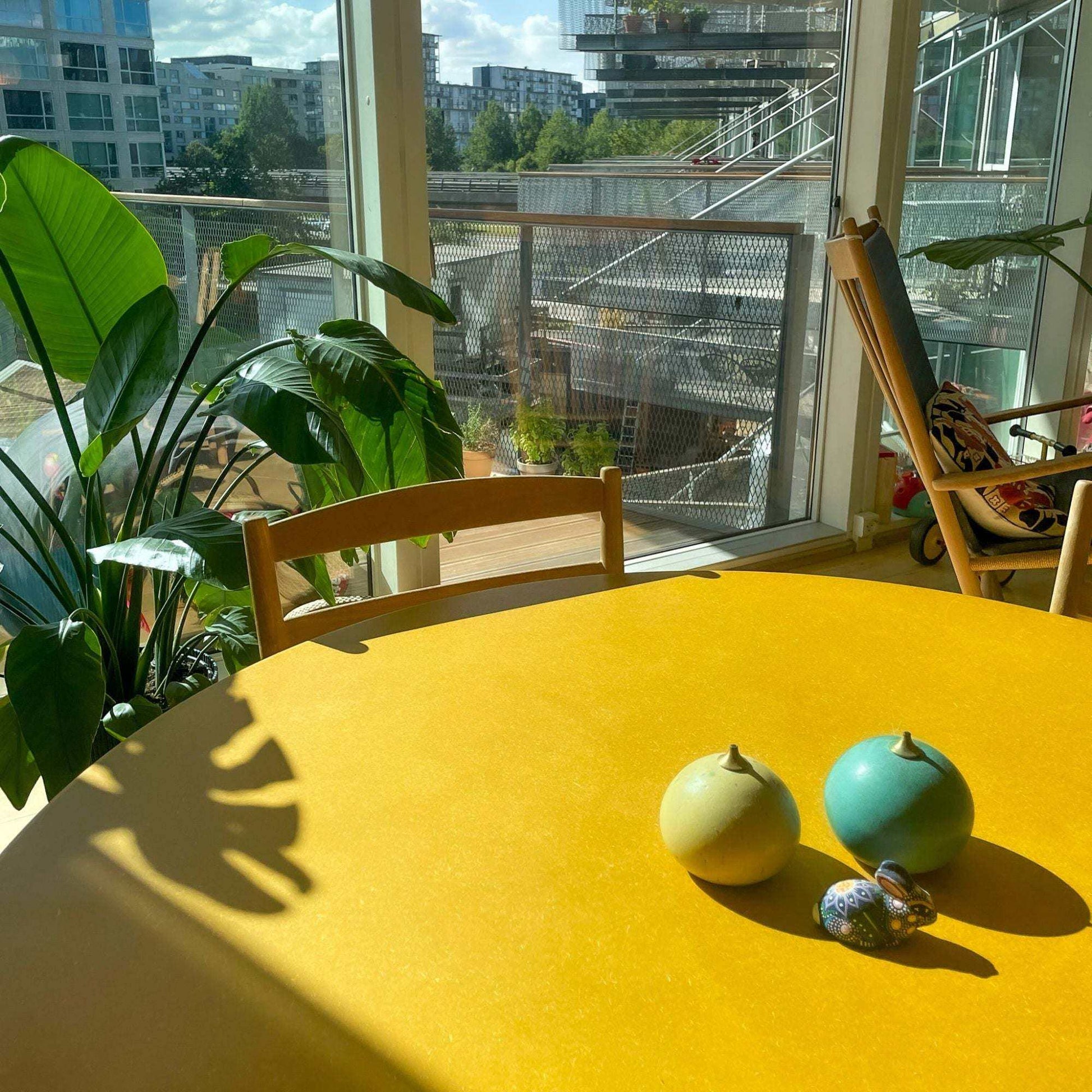 Birch dining table with vibrant yellow Valchromat top in a bright room with plants and balcony view